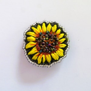 Könnte beinhalten: Gestickte Sonnenblumenbrosche mit gelben Blütenblättern, orangefarbenen Akzenten und einem dunkelbraunen Zentrum. Die Brosche ist mit einem silbernen Perlenrand umrandet, wodurch ein detailliertes Blumenaccessoire entsteht. Die Sonnenblume ist vor einem weißen Hintergrund platziert.