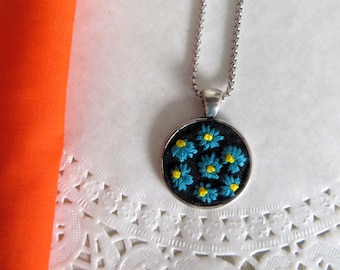 May include: A silver-toned necklace with a round pendant featuring embroidered blue daisies with yellow centers on a black background. The necklace is displayed on a white, patterned surface, with a folded orange fabric in the background.