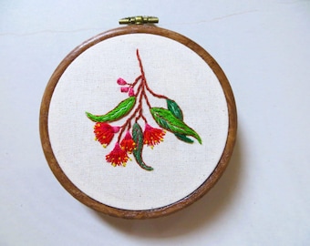May include: Embroidered floral art in a wooden hoop. The design features red and yellow flowers, green leaves, and a brown stem, all stitched on a white fabric. The hoop is approximately 5 inches in diameter.