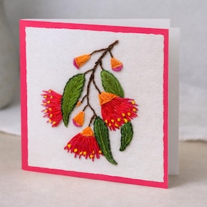 May include: Embroidered greeting card with a floral design. The card has a white background and a pink border. The embroidery depicts red flowers, green leaves, and orange buds. The card is square and folded.