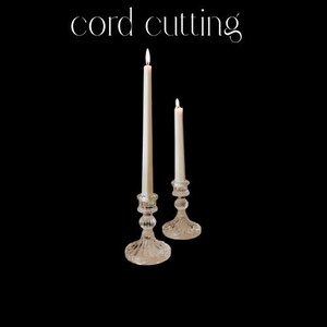 May include: Two lit white candles in ornate glass candle holders against a black background. The taller candle is on the left, and the shorter one is on the right. The text "cord cutting" is at the top.