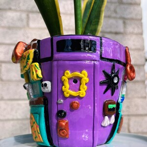 May include: A purple ceramic planter with a painted design of a door with a peephole, a star, and other details. The planter is decorated with a yellow frame around the peephole, and a green and blue design on the side. A green plant is growing out of the planter.