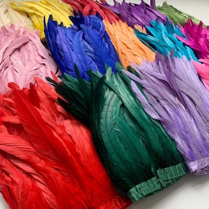 May include: A selection of dyed feathers in a range of colours: red, pink, yellow, blue, green, orange, purple, and turquoise. The feathers are arranged in layers, displaying their vibrant colours and textures. Suitable for craft projects and decorative purposes.