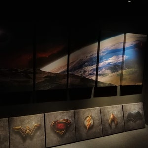 May include: A wall display of six framed images of space scenes, including a planet, a nebula, and a star field. Below the images are nine smaller framed images of superhero logos, including Superman, Wonder Woman, Batman, and The Flash.