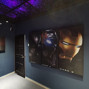 May include: Three movie posters on a blue wall with a black door in the background. The posters are for Iron Man movies, with the text "Mark 1", "Mark 2", and "Mark 3" below each poster. The ceiling is black with purple and white lights.