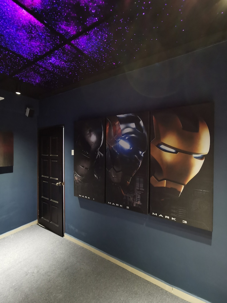 May include: Three movie posters on a blue wall with a black door in the background. The posters are for Iron Man movies, with the text "Mark 1", "Mark 2", and "Mark 3" below each poster. The ceiling is black with purple and white lights.