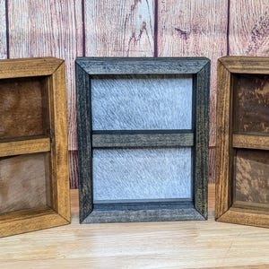 May include: Three wooden framed shadow boxes in varying shades of brown and gray. The center frame is gray, while the other two are brown. Each frame has a textured background.