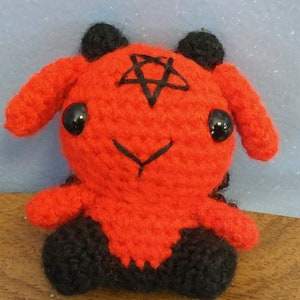 May include: A handmade crochet plushie in the shape of a red creature with black accents. The plushie has a pentagram on its head and large black eyes. The creature has small black horns and is sitting on a wooden surface.