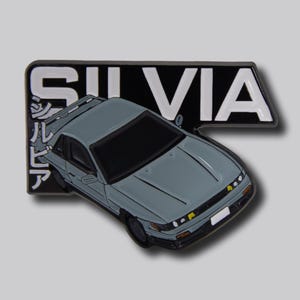 May include: A gray car pin with the word "SILVIA" in white letters on a black background. Japanese characters are on the left side of the pin. The car has a black roof and yellow headlights.