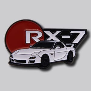 May include: A white RX-7 car pin with black wheels and accents. The pin features the text "RX-7" in black and white, with a red circle behind the "RX" and a white car in the foreground.