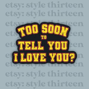 Puede incluir: Pegatina negra con el texto "TOO SOON TO TELL YOU I LOVE YOU?" en letras amarillas con contorno rojo. La pegatina tiene un borde negro y está sobre un fondo gris.