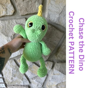 May include: Green crocheted dinosaur toy with yellow horns and black eyes. The dinosaur is being held by a hand.