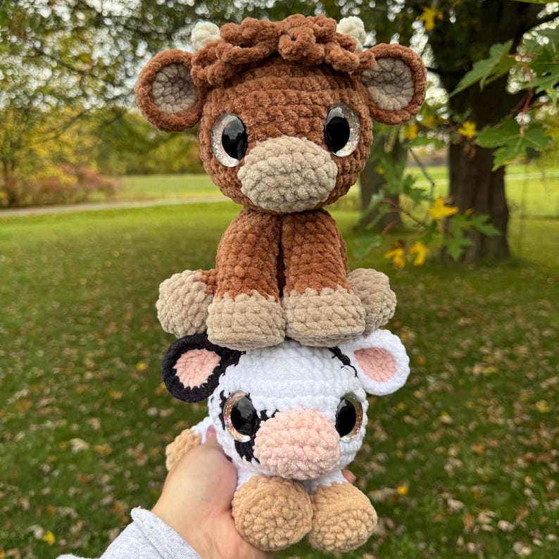 May include: Two handmade crochet cow plushies. The top cow is brown with tan hooves and a light brown muzzle. The bottom cow is white with black spots, pink ears, and tan hooves. Both have large, sparkly eyes.