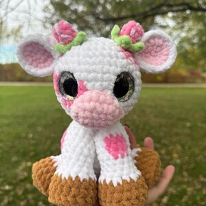 May include: A handmade crochet cow plushie with a white body, pink spots, and a pink nose. The cow has large, glittery eyes and is adorned with pink flower accents on its head. The hooves are brown.