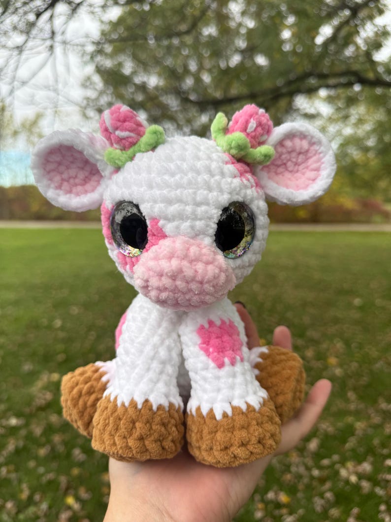 May include: A handmade crochet cow plushie with a white body, pink spots, and a pink nose. The cow has large, glittery eyes and is adorned with pink flower accents on its head. The hooves are brown.