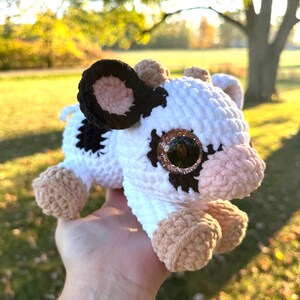 May include: A handmade crochet cow plushie with a white body, black spots, and brown accents. The cow has large, glittery eyes and is held in a hand. The word "Believe" is tattooed on the person's wrist.