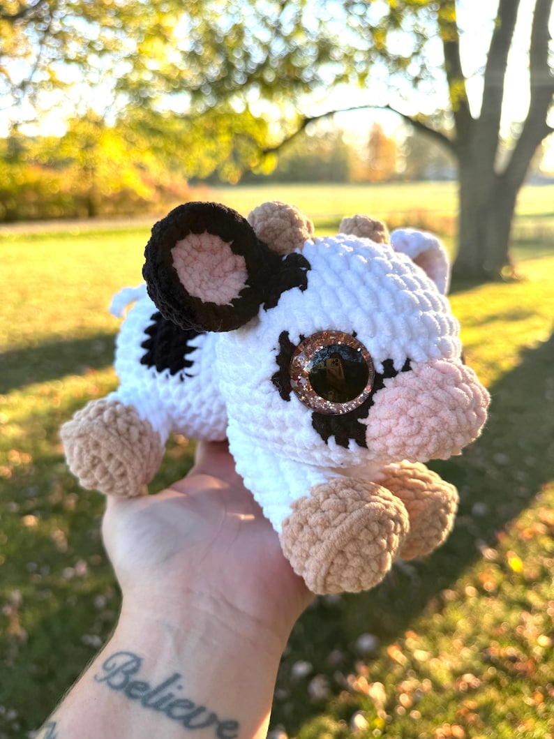 May include: A handmade crochet cow plushie with a white body, black spots, and brown accents. The cow has large, glittery eyes and is held in a hand. The word "Believe" is tattooed on the person's wrist.