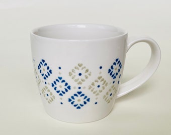 May include: White ceramic mug with a curved handle. The mug features a repeating pattern of blue and silver geometric shapes around the body. The design is a series of diamond and dot shapes. The mug is on a white background.