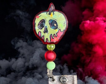 May include: A green and red apple-shaped badge holder with a skull and crossbones design. The holder is attached to a beaded chain with red and green beads. The holder is against a black and red smoke background.