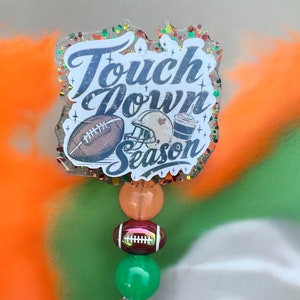 May include: A clear badge holder with a football, helmet, and drink design. The design says "Touch Down Season". The badge holder has orange, green, and brown beads.