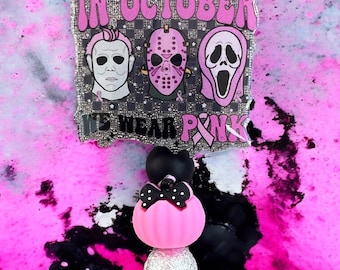May include: A Halloween-themed keychain with a pink pumpkin, a black bow, and a silver glitter bead. The top features a banner with the text "IN OCTOBER WE WEAR PINK" and images of horror movie characters.