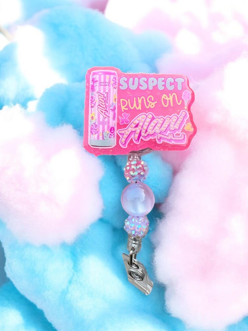 Pu&ograve; includere: Un porta badge rosa con la scritta "SUSPECT runs on Alani!" presenta una grafica di lattina di bevanda energetica Alani Nu rosa. Il porta badge ha un accento di perline con una clip argentata. Lo sfondo &egrave; zucchero filato blu e rosa.