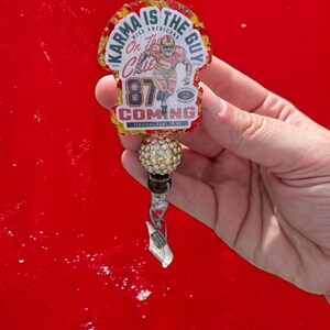 May include: A red, gold and white charm with the text "Karma is the Guy" and "On the Chic 87 Coming Straight Home to Me". The charm features a cartoon image of a football player wearing a red jersey with the number 87. The charm is attached to a silver clip and a beaded chain with a crystal-encrusted bead.