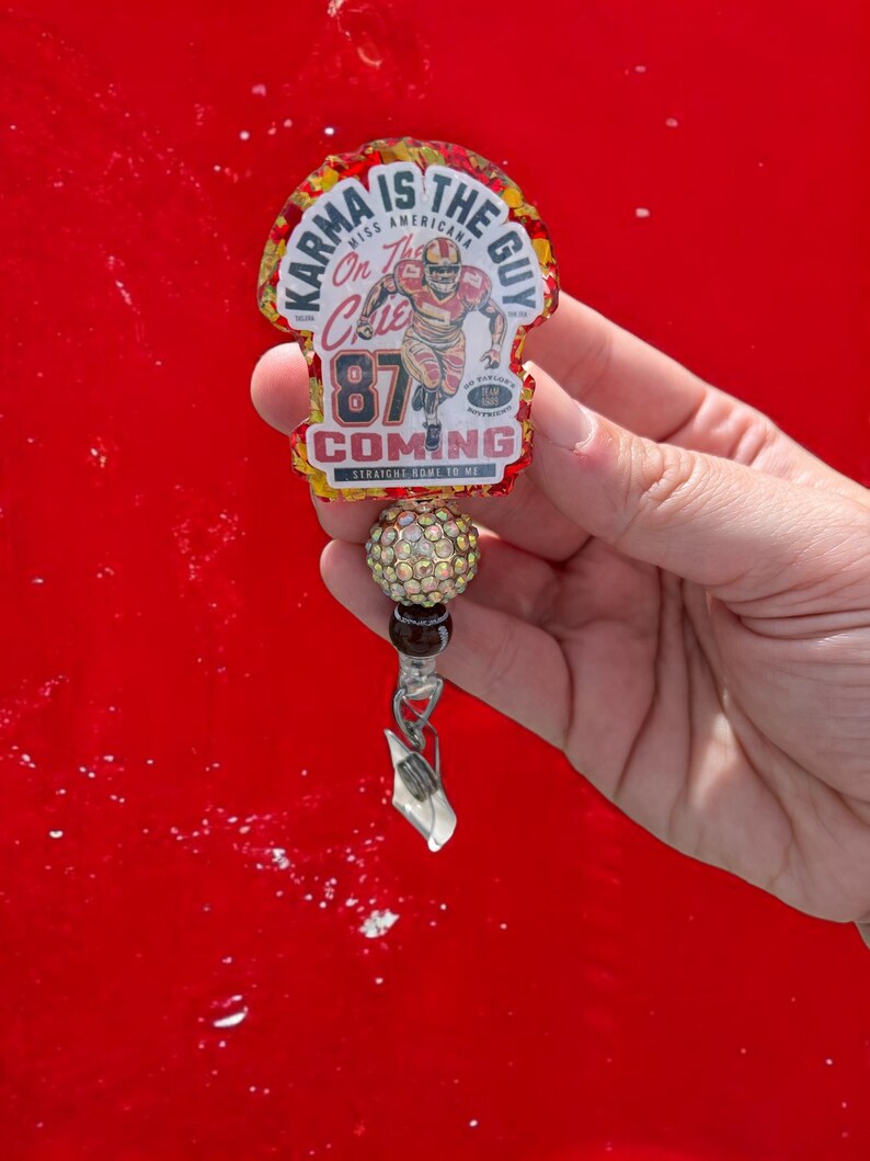 May include: A red, gold and white charm with the text "Karma is the Guy" and "On the Chic 87 Coming Straight Home to Me". The charm features a cartoon image of a football player wearing a red jersey with the number 87. The charm is attached to a silver clip and a beaded chain with a crystal-encrusted bead.