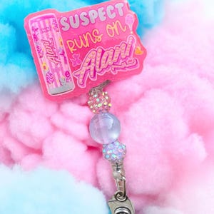 Pu&ograve; includere: Un porta badge rosa con la scritta "SUSPECT runs on Alarm!" e una grafica di un contenitore di zucchero filato rosa. Il porta badge ha un accento di perline e una clip argentata. Lo sfondo &egrave; rosa e blu.