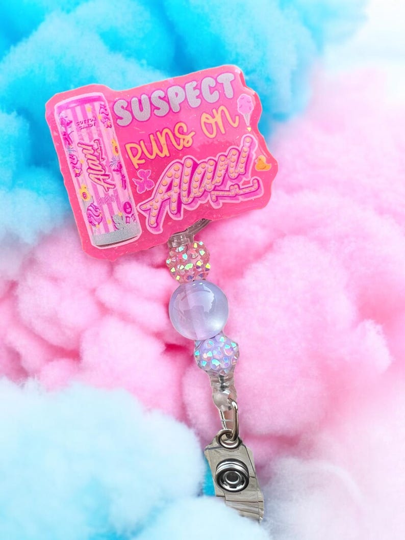 Pu&ograve; includere: Un porta badge rosa con la scritta "SUSPECT runs on Alarm!" e una grafica di un contenitore di zucchero filato rosa. Il porta badge ha un accento di perline e una clip argentata. Lo sfondo &egrave; rosa e blu.