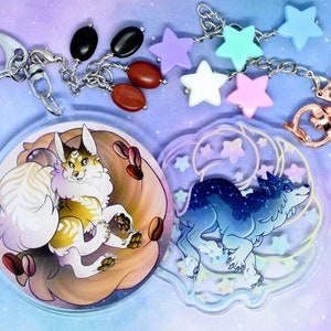 May include: Two round clear acrylic charms with a keychain attached. One charm features a white and orange cat with brown coffee beans. The other charm features a blue wolf with a crescent moon and stars. The keychain has a silver chain with a silver ring and a gold star charm.