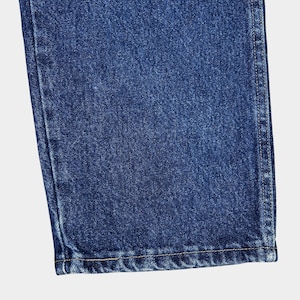 Pu&ograve; includere: Primo piano del bordo inferiore di un paio di jeans blu denim. Il tessuto ha un aspetto testurizzato e leggermente sbiadito. L'orlo &egrave; cucito con un filo di colore chiaro.