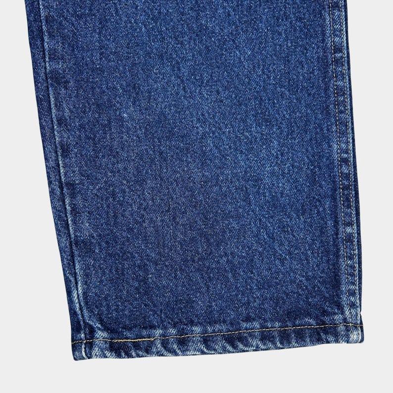 Pu&ograve; includere: Primo piano del bordo inferiore di un paio di jeans blu denim. Il tessuto ha un aspetto testurizzato e leggermente sbiadito. L'orlo &egrave; cucito con un filo di colore chiaro.