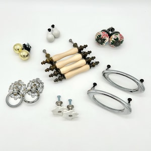 May include: A collection of assorted cabinet hardware including four cream-colored bar pulls with brass-toned ends, two silver lion head ring pulls, two floral ceramic knobs, two gold-toned spherical knobs, two white knobs, and two oval silver pulls.