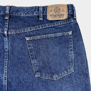 Pu&ograve; includere: Un paio di jeans blu denim con una tasca posteriore e una toppa in pelle marrone che dice "Wrangler Authentic Jeans" e "Premium Quality".