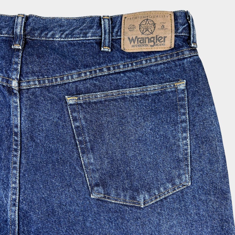 Pu&ograve; includere: Un paio di jeans blu denim con una tasca posteriore e una toppa in pelle marrone che dice "Wrangler Authentic Jeans" e "Premium Quality".