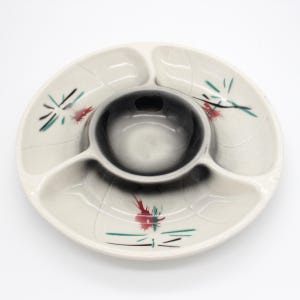 Op de afbeelding: Een vintage, crèmekleurige keramische serveerschaal met een centrale kom en drie omliggende compartimenten. De schaal heeft een zwart-grijze gradiënt in het midden en is versierd met rode, zwarte en turquoise bloemaccenten. Een stijlvol stuk voor het serveren van snacks of hapjes.