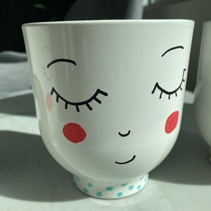 May include: White ceramic planter with a hand-painted face, closed eyes, and a small smile. The planter has red cheeks and a blue and white polka dot pattern around the bottom.