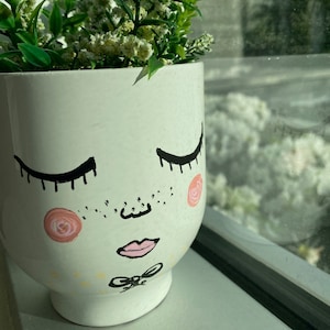 May include: White ceramic planter with a hand-painted face, featuring closed eyes, a nose, and a pink mouth. The planter has pink and white floral accents and is filled with green plants.
