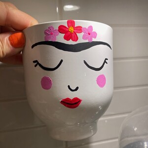 Può includere: Vaso in ceramica bianca con un viso dipinto, con occhi chiusi, un piccolo sorriso e labbra rosse. Il vaso è decorato con fiori rosa e rossi.