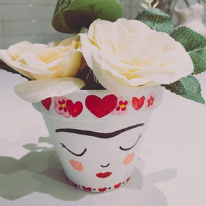 Puede incluir: Una maceta pintada de blanco con una cara pintada, ojos cerrados, labios rojos y rubor. El borde está decorado con corazones rojos y flores rosas. La maceta contiene rosas color crema y follaje verde.