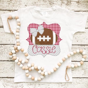 Puede incluir: Camiseta blanca con un marco a cuadros rojo y blanco, una pelota de fútbol marrón con un lazo blanco y el nombre "Cassie" en rayas grises y blancas.