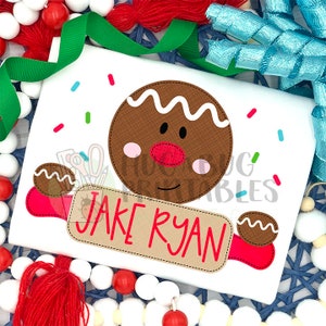 May include: A white t-shirt featuring a gingerbread man design. The gingerbread man is holding a rolling pin with the text "JAKE RYAN" on it. The design is surrounded by colourful sprinkles.