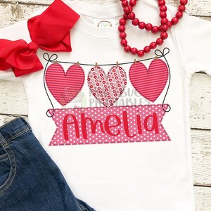 May include: A white t-shirt with a pink banner that says "Amelia" and three red hearts with different patterns hanging from a string.