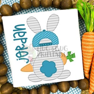 May include: A white bunny with gray stripes wearing a blue and white checkered hat and holding a carrot. The bunny is holding the carrot with its right paw. The text "Happy Easter" is written in black on the white background.