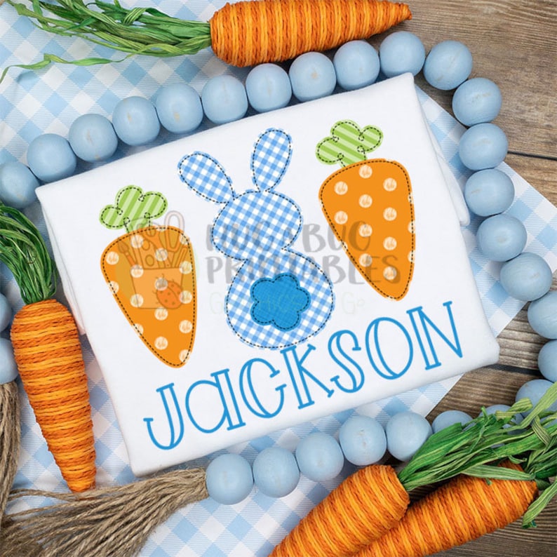 May include: A white onesie with a blue bunny, two orange carrots, and the name "Jackson" embroidered in blue. The bunny has a blue flower on its chest. The carrots have green tops and white polka dots.