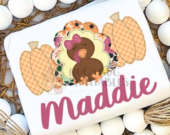 May include: A white onesie with a cute embroidered turkey design. The turkey is surrounded by fall-colored flowers and pumpkins. The onesie is personalized with the name "Maddie" in pink.