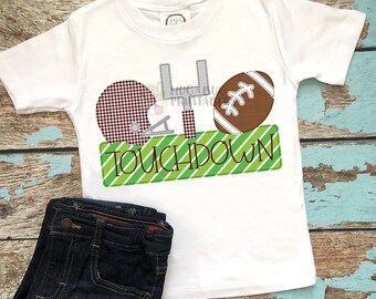 May include: White t-shirt featuring a brown football, a red and white checked football helmet, and a grey football goal post. The shirt has a green and white striped banner with the word "TOUCHDOWN" on it.