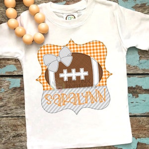 May include: White t-shirt with an orange and white checkered frame with a brown football with a white bow on top. The name "Saralynn" is written in gray letters below the football.