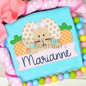 May include: A light blue t-shirt with an embroidered design of a brown bunny rabbit with a pink bow, peeking over a pink and white banner with the name "Marianne" embroidered in blue.